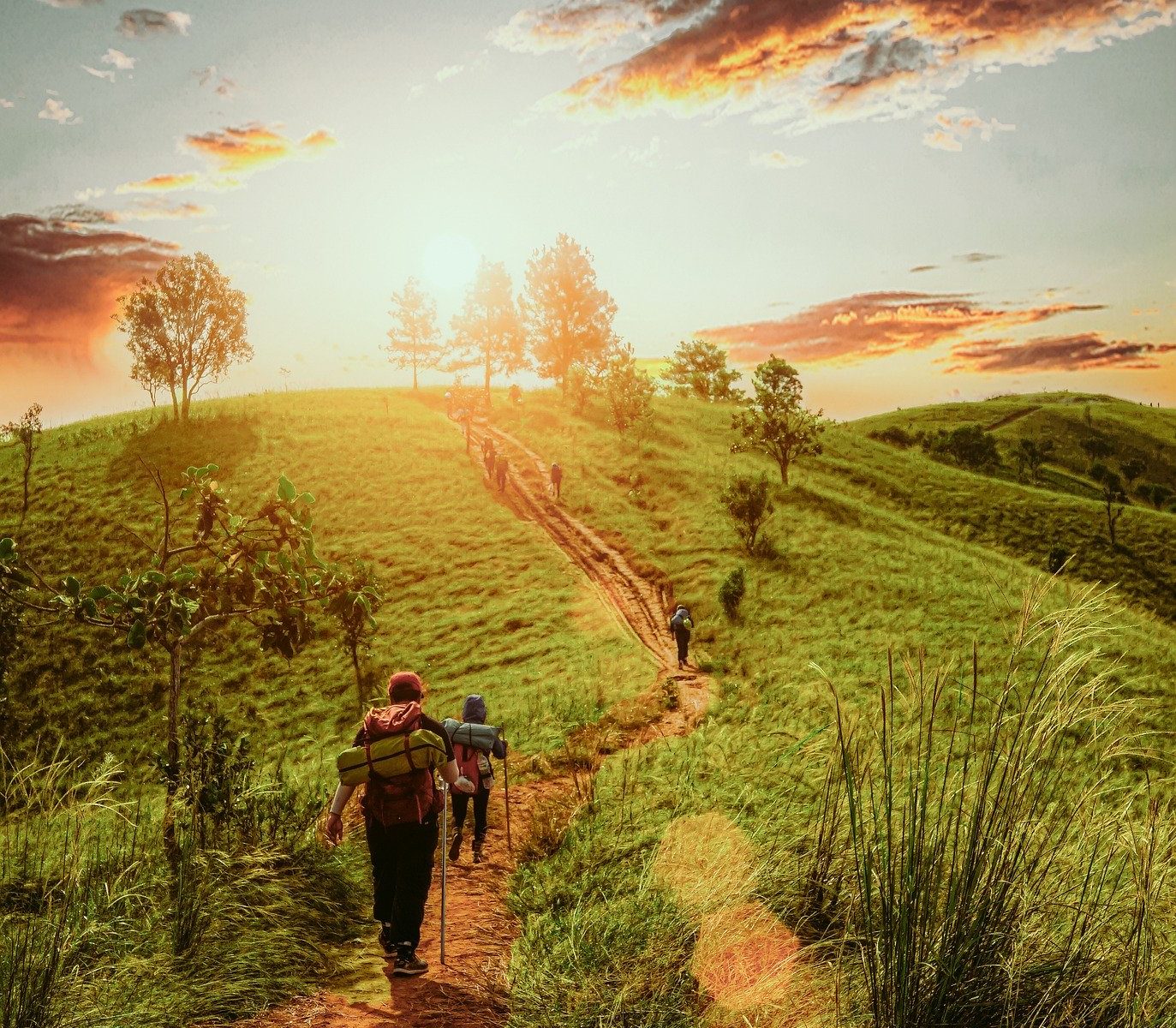 Wanderer auf einem Pfad durchs Grüne, im Hintergrund ein Hügel und die untergehende Sonne